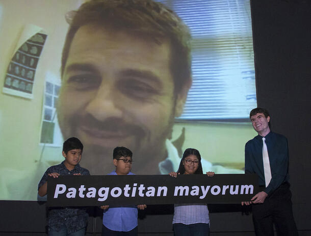 Four people hold a sign reading Patagotitan mayorum while Diego Pol smiles on a Skype call displayed on a large screen behind them.