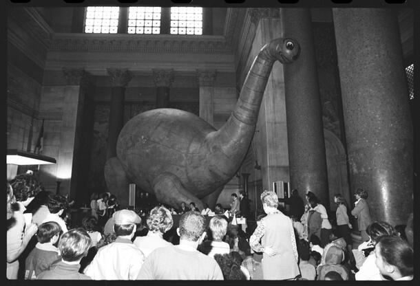 Large crowd stands around inflated dino in the Museum's high-ceilinged rotunda.