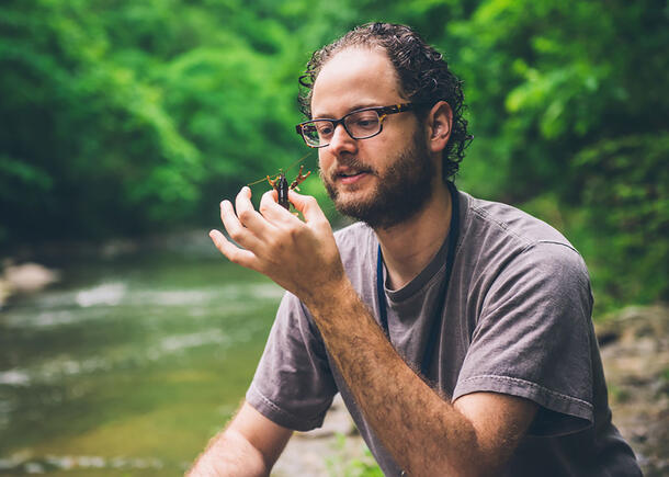 Michael squats near the edge of a stream and holds up a crayfish.