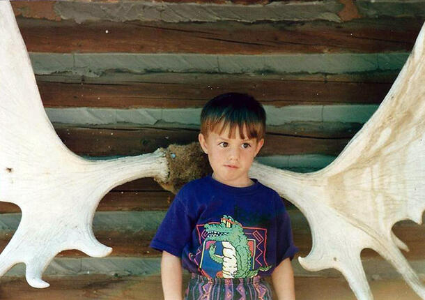 Young Zac stands in the middle of a massive pair of antlers that are mounted on a log cabin wall.