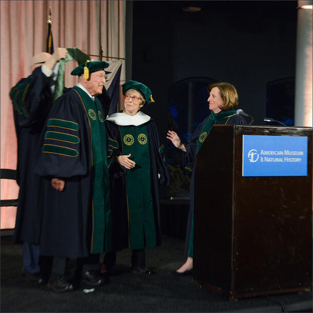A graduation cap is places on a man's head as he stands near a podium.