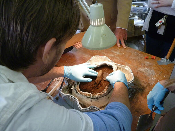 People gather close to a table to watch a scientist ease a piece of copper from an earth sample in a foil container.
