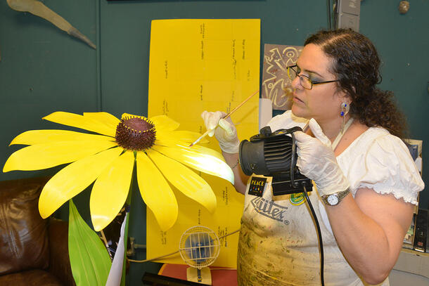 Woman paints the petal on a larger-than-life flower with one hand while using her other hand to direct a uv light at the fresh paint.