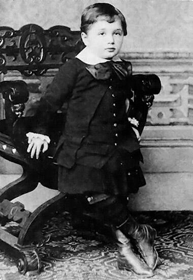 Young Albert Einstein stands leaning against a carved, wooden chair.