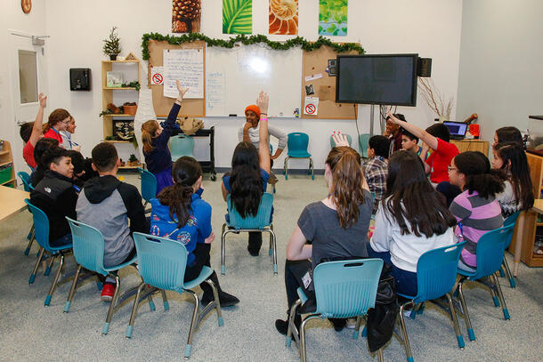 A group of students sit in chairs grouped in a semi-circle around their instructor and raise their hands in response to a question.