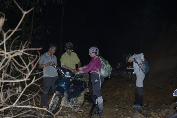 Four adults standing outdoors on brown rocky dirt near a motorcycle. Captions reads: “Preparing our equipment before our night survey in Bach Ma National Park.”