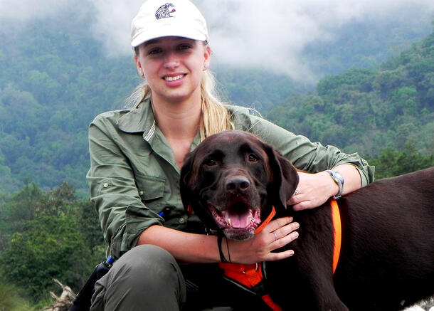 Person in a baseball cap with long, light hair sits with arms around a dark-colored, medium-sized dog in front of view of green, misty mountains.