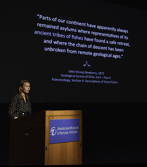 Allison Bronson speaks from a podium, text is visible on the screen behind her and describes the evolution of fossil fishes.
