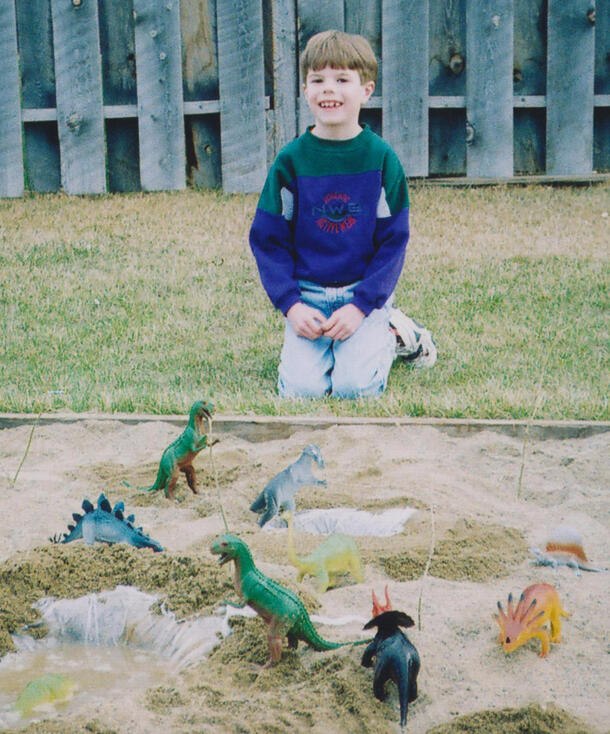 Childhood photo of Danny Barta kneeling next to a sand pit where has posed an assortment of plastic dinosaurs.