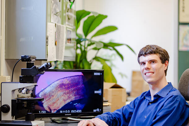 Danny Barta sits at a desk and uses a microscope and computer.