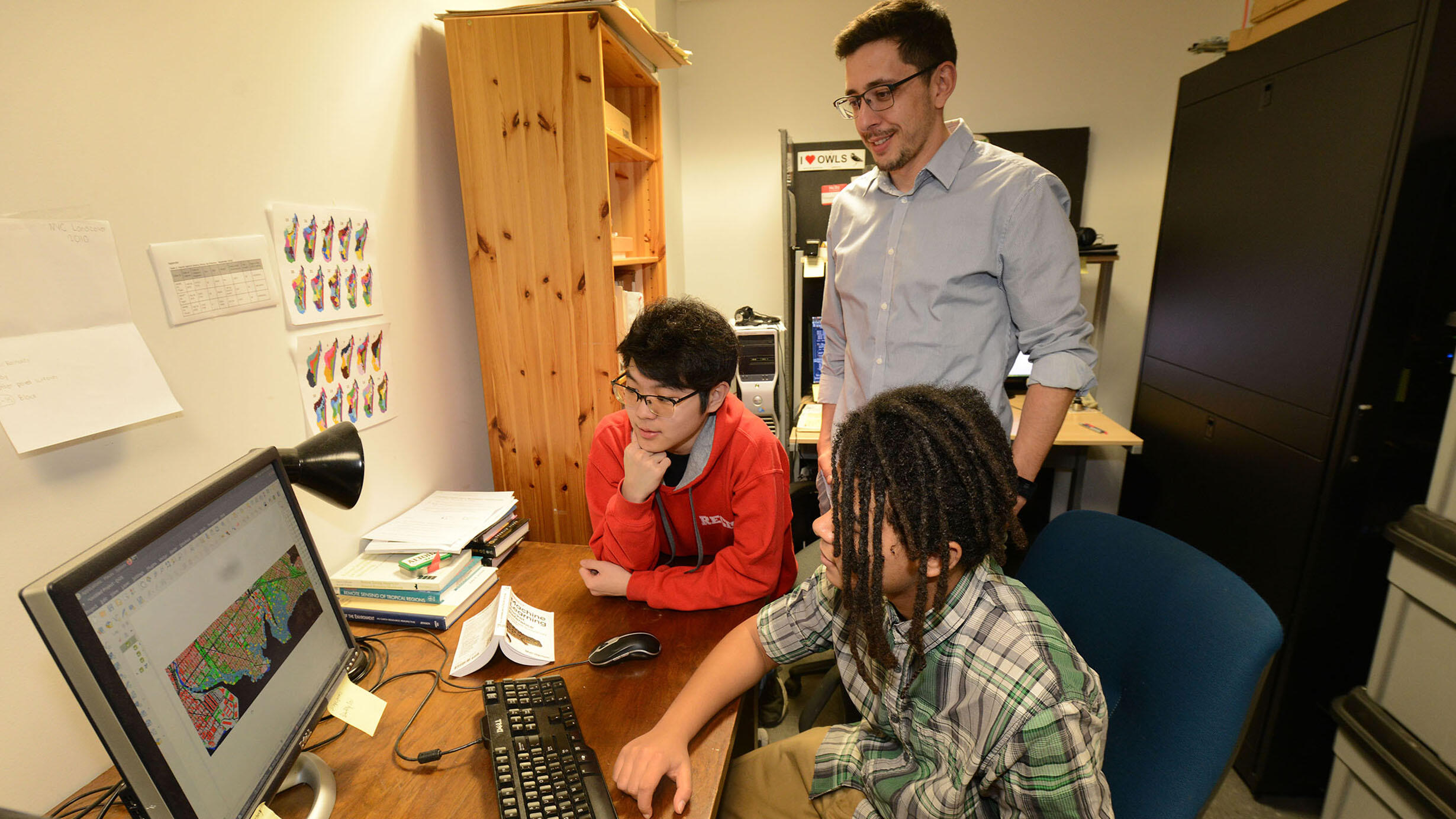 Two students sit at desk and look at a computer monitor while their mentor stands behind them.