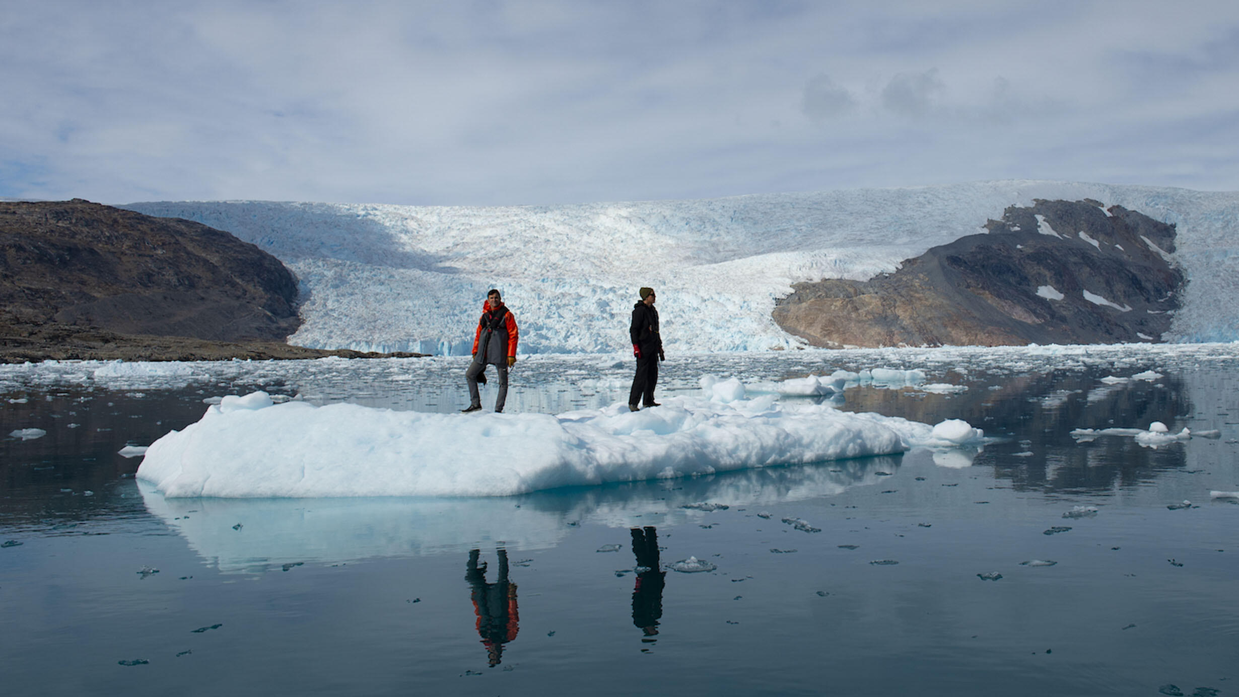 Two scientists in winter gear stand on an iceberg outcrop.