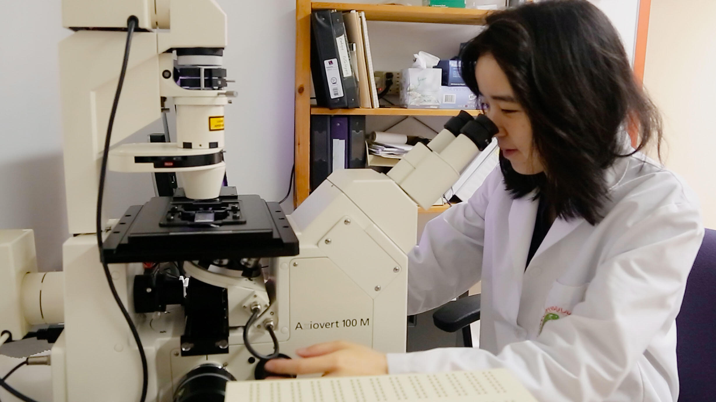 Scientist Eunsoo Kim sits at a lab table, wearing a lab coat and looking into a microscope.