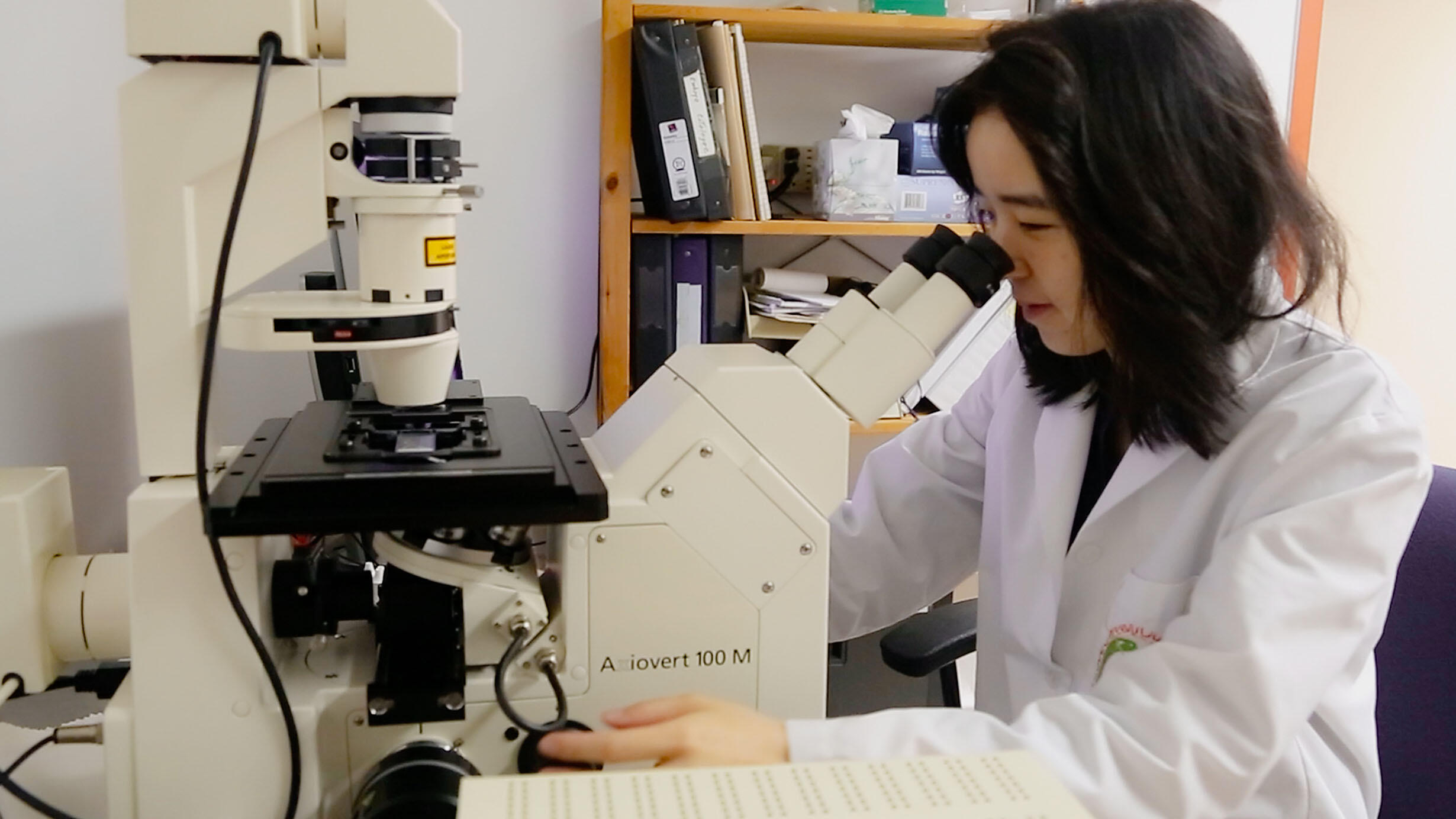 Scientist Eunsoo Kim sits at a lab table, wearing a lab coat and looking into a microscope.