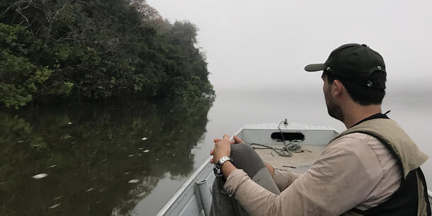 Person facing away from camera sits in a boat, with shoreline on the left.