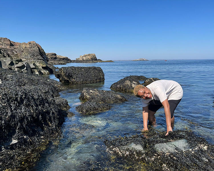 Leanne Melbourne stands in the ocean amidst rocks, close to the shore, and bends down with both hands in the water.