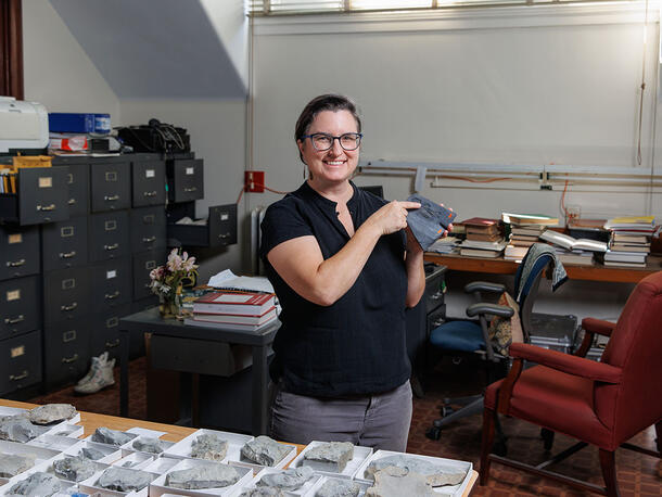 Melanie Hopkins stands in an office with file cabinets and a table covered with fossil specimens and holds a trilobite fossil.