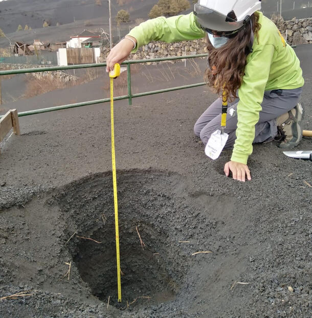 Samantha Tramontano, wearing a helmet and a mask, kneels beside a hole and measures accumulated volcanic ash with a tape measure.