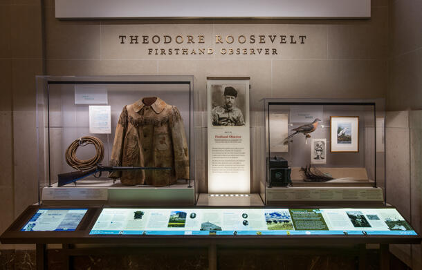 Display case titled "Theodore Roosevelt Firsthand Observer," containing jacket, camera, rifle, bird, and other artifacts with photos and text