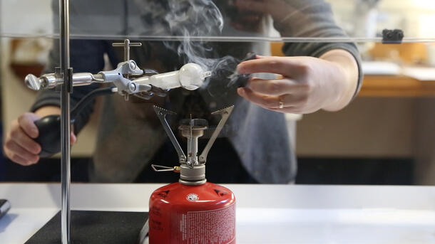 Person at a laboratory table with a small object in one hand, treating it with a gaseous substance coming from a glass tube attached to a small hose with a bulb at the end.