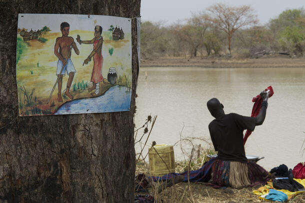 A person seated near water’s edge washing clothes in a large vessel. Nearby is a poster on a tree depicting two people infected with Guinea worm.