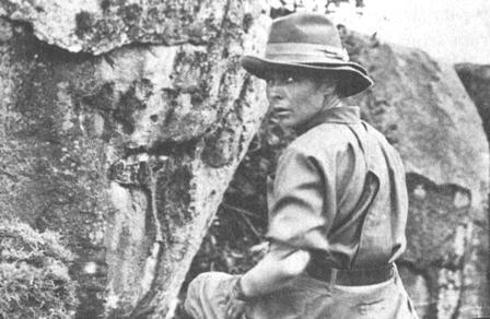 Person outdoors beside rock face in daytime. Looking over her left shoulder backward at camera. Wearing wide brimmed hat and long-sleeved field shirt.
