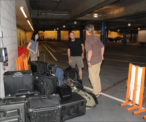 A woman and two men standing in a underground parking lot with many luggage cases.