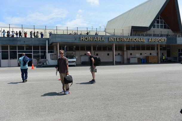 Three people standing on an asphalt street outside a low building with the words, “Honiara International Airport.”