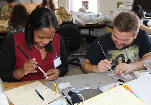 A man and woman seated at a table. Both are smiling as they work with dry paint brushes to clean very small artifact pieces. 