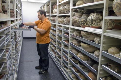 Man standing between two tall rows of shelves holding pottery artifacts. The man has gloves on and has pulled open a tray of smaller artifacts.