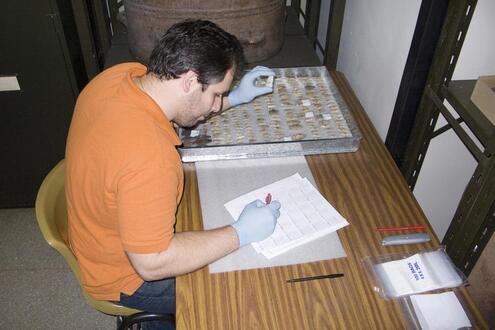 Man seated at a table with a small case of objects. He holds up an artifact with one gloved hand while filling out a form with the other. 