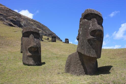 Two giant head scultures on a grassy hill; One head appears partially buried. 