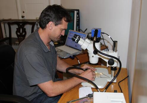 Man in an office seated at a desk, using a microscope to inspect a piece of pottery. In the background, there is a laptop on a nearby desk.