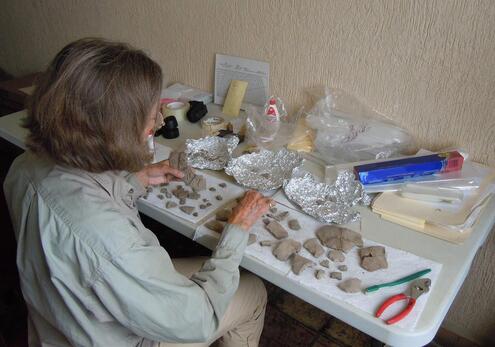 Older woman sorting and piecing together many pieces of pottery spread out on a folding table, along with glue, tin foil, paper and various tools. 