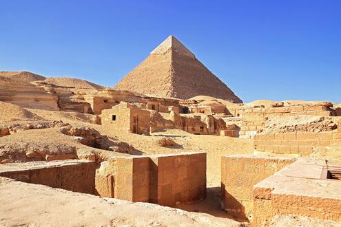 Dusty ruins of simple brick homes (many missing their roofs) with large pyramid looming in the background against a clear blue sky. 