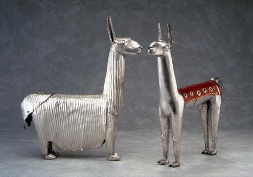 Two small llama figurines made of silver. One is stylized with a long, full coat, and the other appears shaven, with a colored blanket on its back.