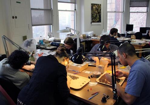 Five people seated at a lab table, working under the light of several swing-arm lamps to sift through trays of dirt for artifacts.