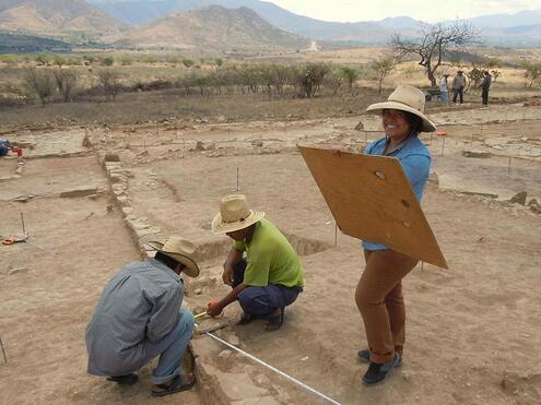 Woman holding a large wooden drawing board, next to two men kneeling down with a measuring tape surveying an area. 