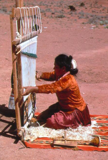 Woman kneeling in front of a large wooden loom weaving a textile.