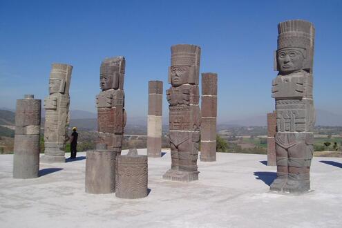 A line of four towering warrior statues. Several pillars surround them, and a small man can be spotted in the background. 