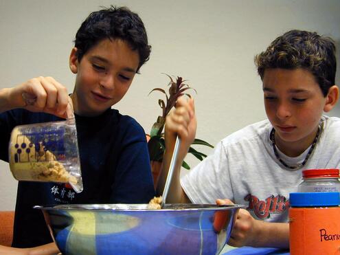 Two boys over a large metal mixing bowl; one boy is pouring wheat germ into the bowl while the other stirs the bowl's mixture. 