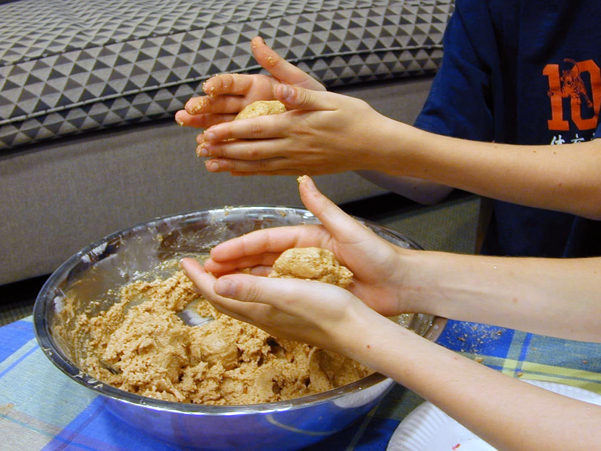 Closeup of two pairs of boys' hands rolling cookie dough into balls before a large metal bowl.