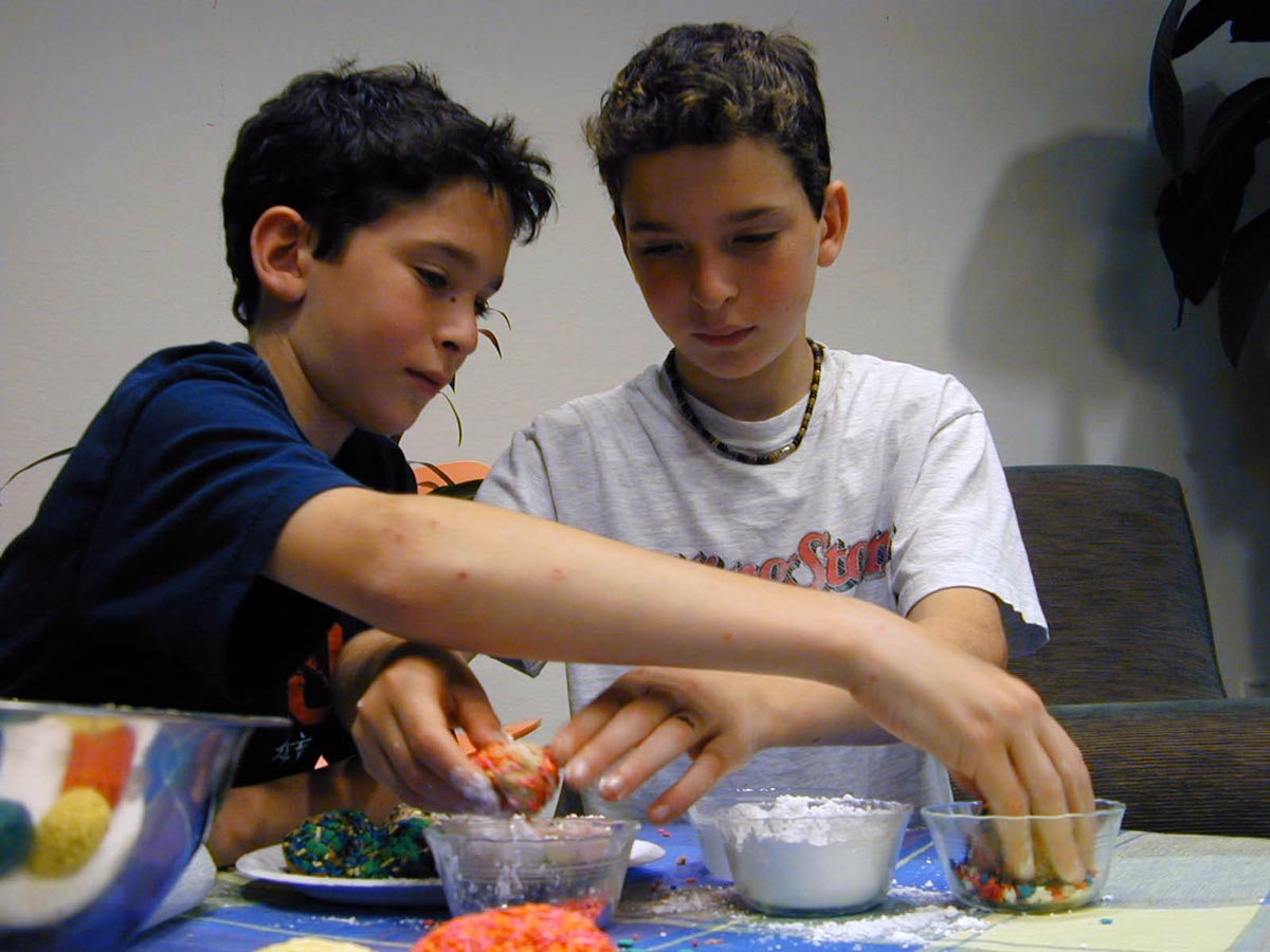 Two boys rolling cookie dough balls in containers of sprinkles and powdered sugar.