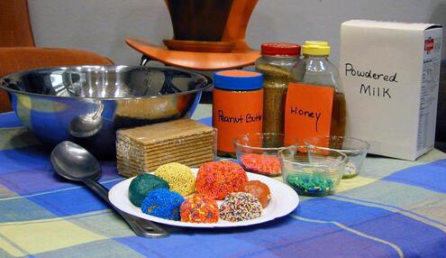 Table with peanut butter, honey, powdered milk, graham crackers, a mixing bowl and serving spoon, sprinkles and several decorated cookie balls. 