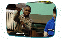 Two children sit next to each other. The child to the left holds a ball of twine and prepares to toss the twine at someone while holding the string.