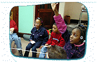 Five seated children playing a game involving a piece of string, with one child raising their hand.