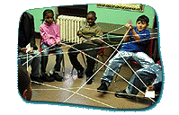 Children seated in a circle hold different corners of a long piece of string to create triangular, overlapping shapes between them.