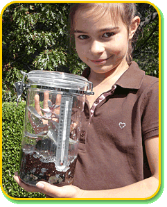A child outdoors smiling at the camera and holding a clear cylindrical container with a plant and a thermometer.