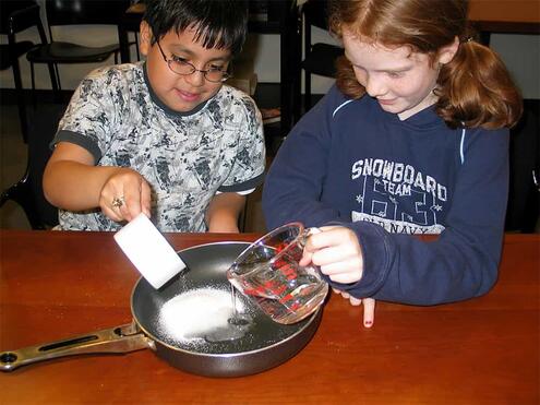 one child pouring sugar into a saucepan while the other child pours in water