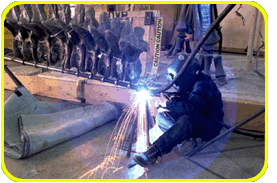 A person in a welding mask sits and welds a large piece of metal, which will be part of the spinal support on the skeletal mount for a T. Rex.